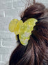 Yellow butterfly hair clip in a person's hair against a light gray brick wall.