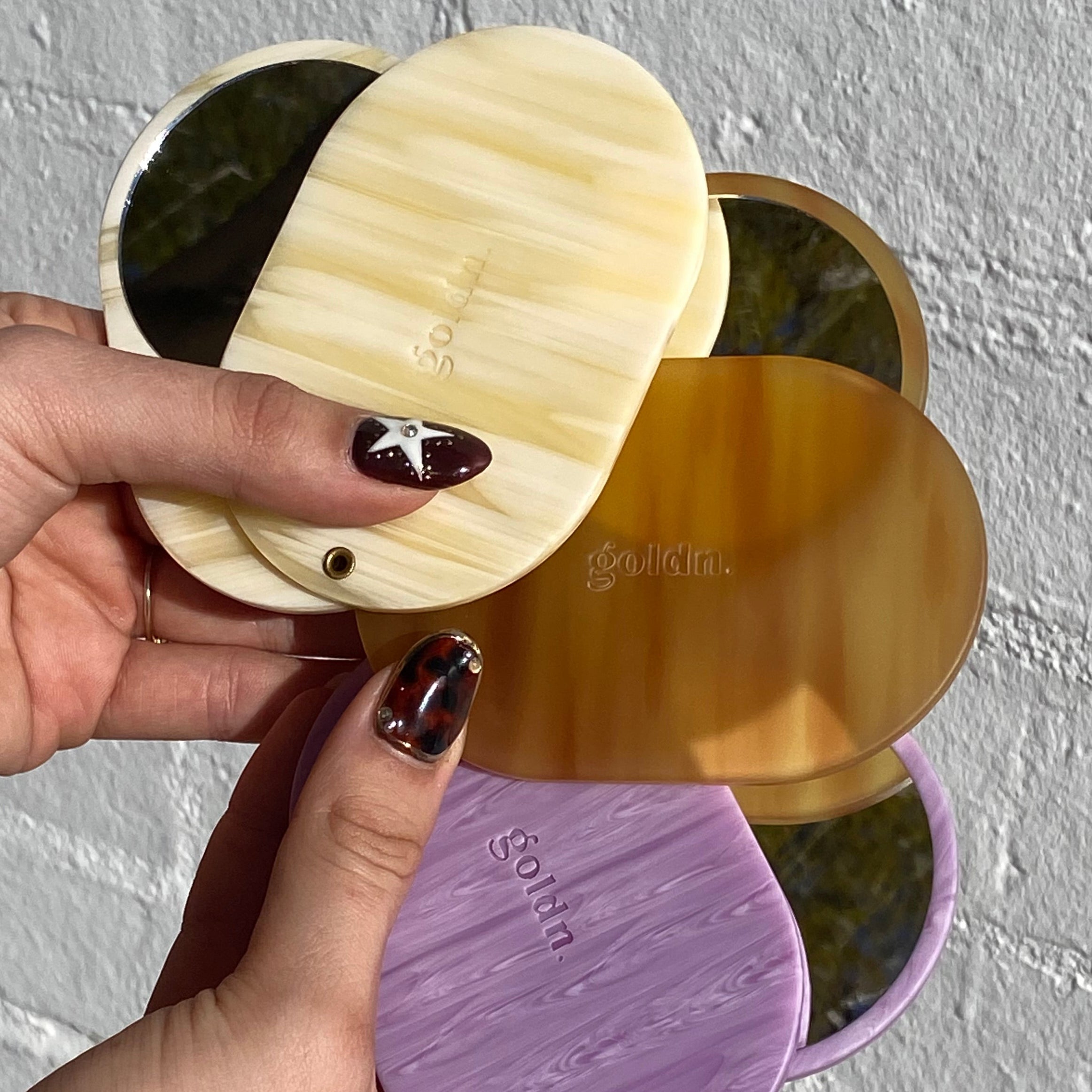 Hand holding four round, reusable makeup pads in gold, beige, and purple against a textured gray background.