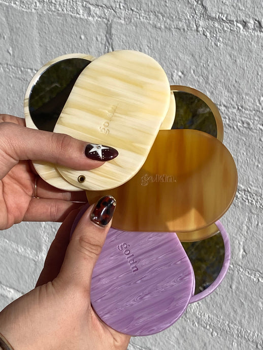 Hand holding four round, reusable makeup pads in gold, beige, and purple against a textured gray background.