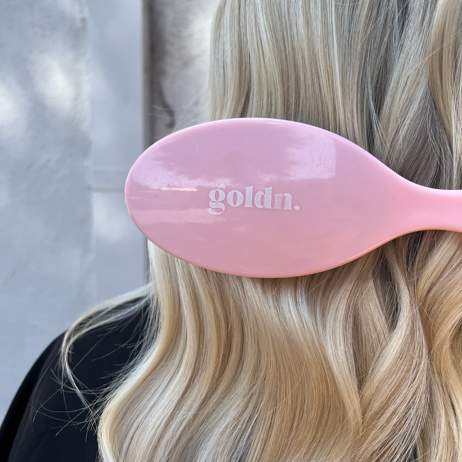 Pink hairbrush with 'goldn.' branding held by a person with blonde hair.