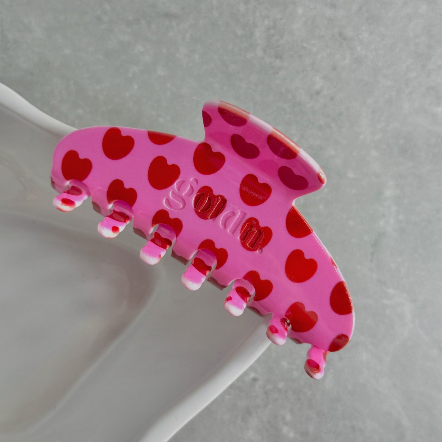 Pink hair clip with red heart patterns on a gray surface. Goldnwares Curve Clip with a heart pattern plate-based acetate hair clip for medium to thick hair.