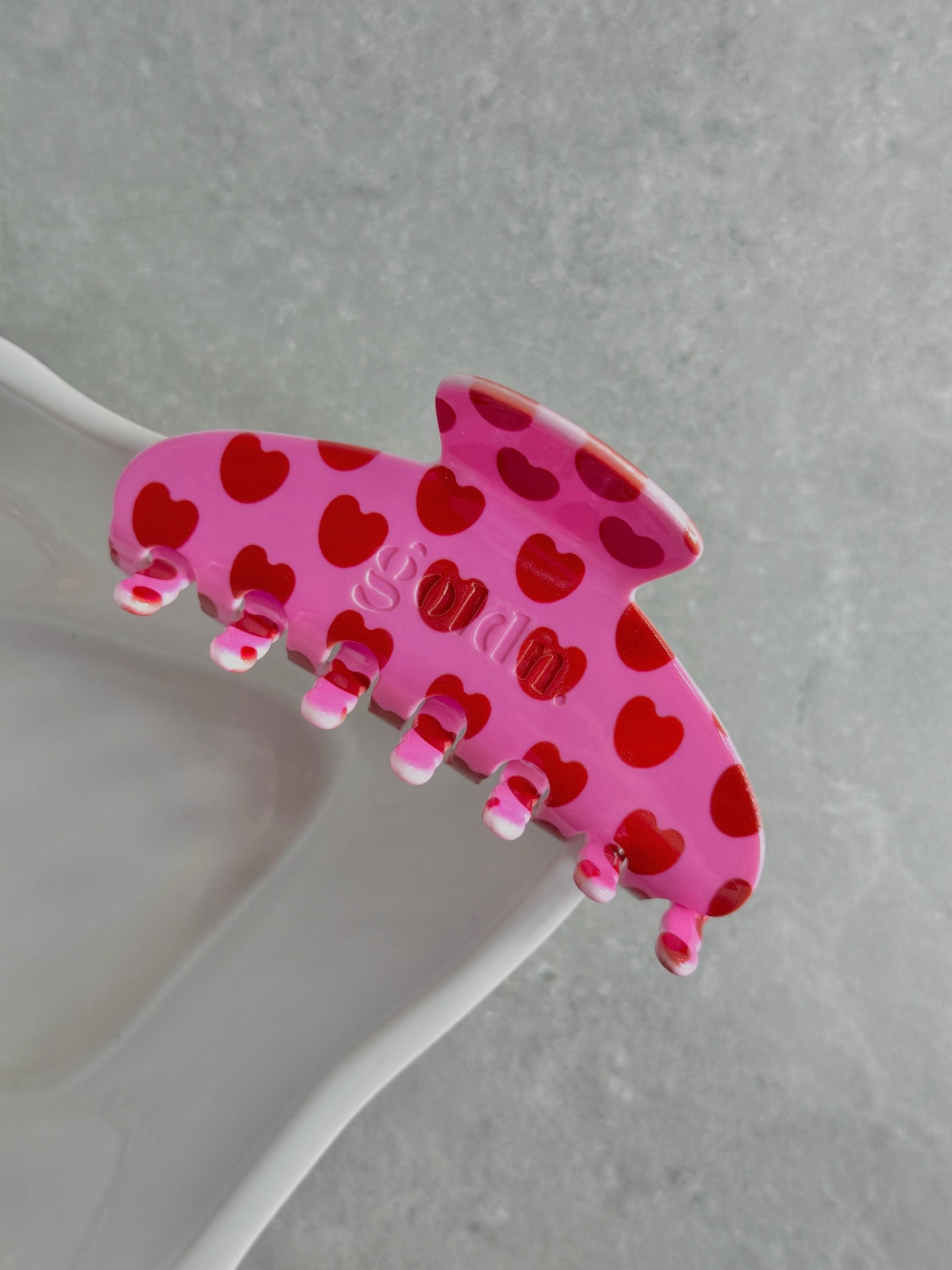 Pink hair clip with red heart patterns on a gray surface. Goldnwares Curve Clip with a heart pattern plate-based acetate hair clip for medium to thick hair.