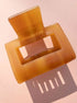 Translucent amber Lounge Claw Clip Small with rectangular cutout, casting shadow on pink surface for aesthetic appeal.