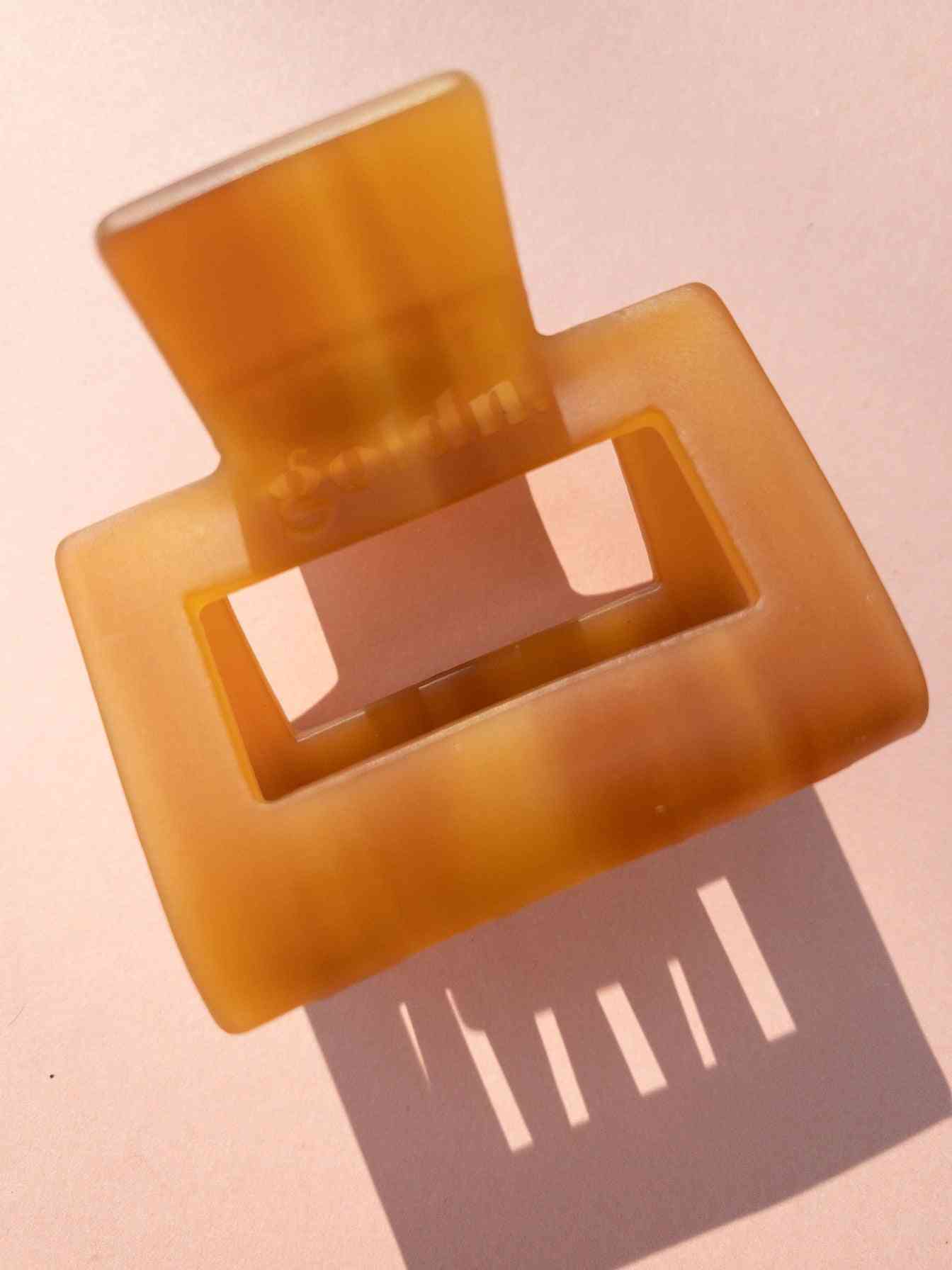 Translucent amber Lounge Claw Clip Small with rectangular cutout, casting shadow on pink surface for aesthetic appeal.